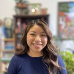 Jenny has long, wavy hair with blonde highlights. She is wearing a navy blue shirt. She is smiling.